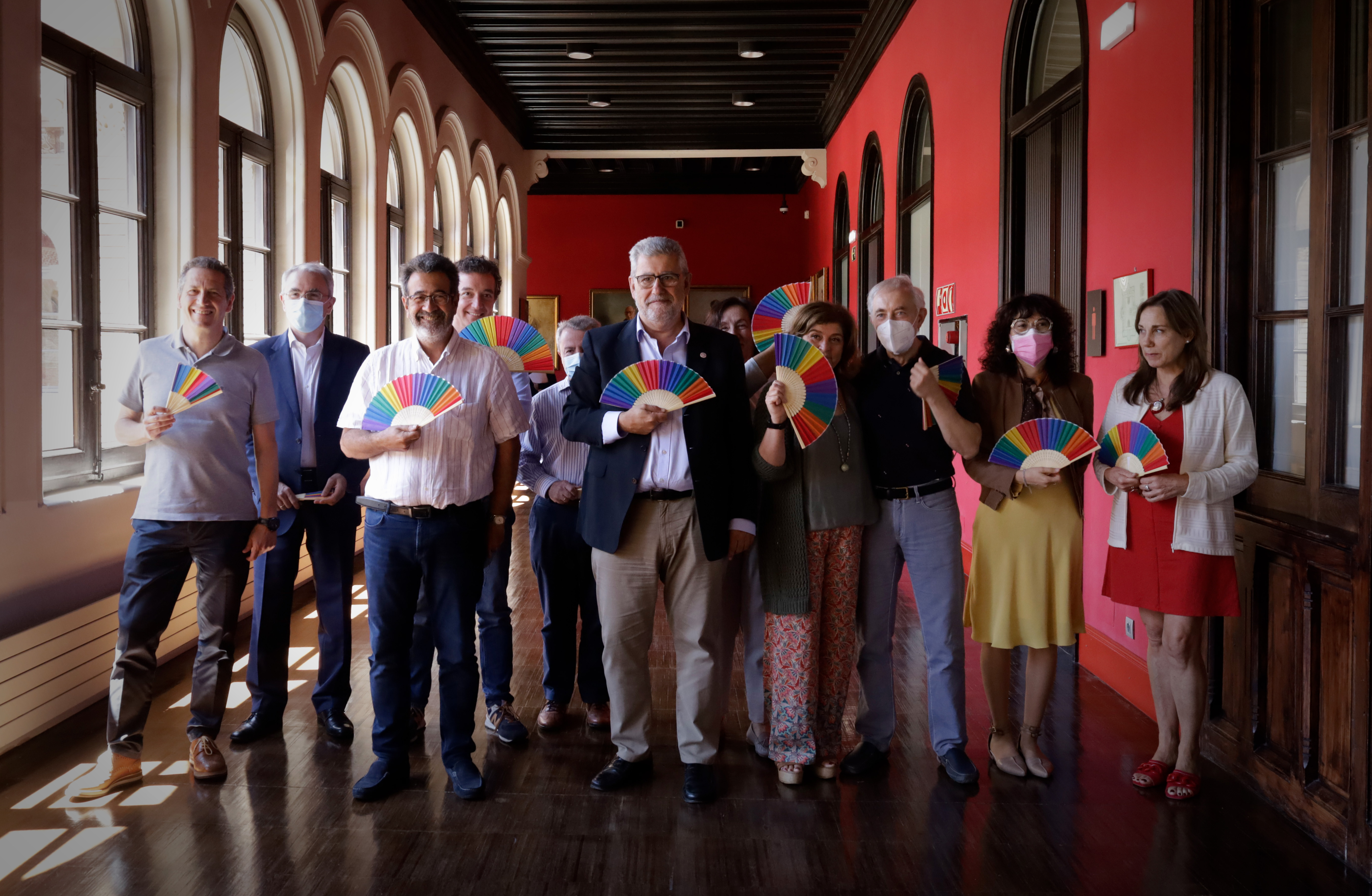 Foto del consejo de gobierno de la Universidad de Zaragoza portando abanicos con los colores representativos de la bandera arcoiris LGBT+ en el pasillo del Paranifo